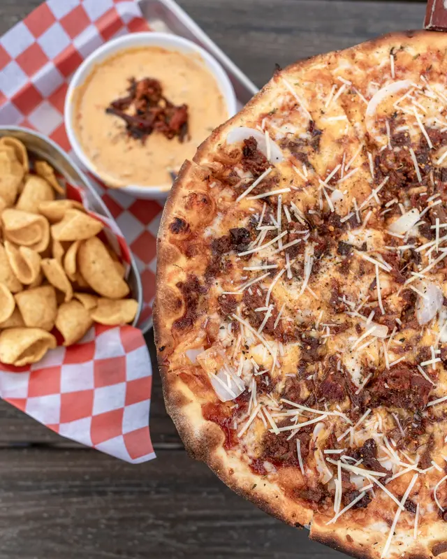 Wood-fired pizza topped with shredded meat and cheese, served with cashews and a creamy dipping sauce on checkered paper